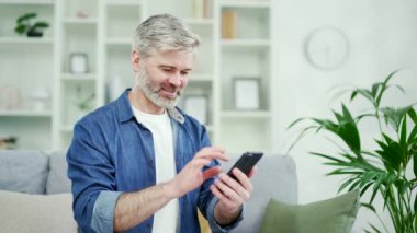 Gülümseyen, gri sakallı, akıllı telefon kullanan, evdeki koltukta otururken mesaj okuyan bir adam. Mutlu adam sosyal ağları tarıyor, SMS yazıyor, e-postaları kontrol ediyor ya da internette sohbet ediyor.