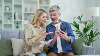 Mutlu evli çift, evdeki kanepede otururken online fatura ödeme uygulamasını kullanarak aile sermayesiyle ilgileniyor. Olgun gülümseyen eş elinde çekler tutuyor ve kocası akıllı telefondan verileri kaydediyor.
