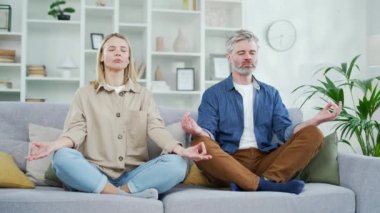 Evde oturma odasında kanepede otururken nilüfer pozisyonunda gözleri kapalı meditasyon yapan olgun evli bir çift. Karı koca, rahat bir kanepede sakince nefes alırken yoga yapıyorlar.