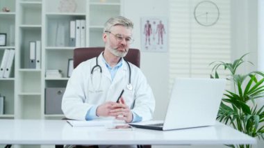 Hastane kliniğinde otururken bilgisayarla konuşan beyaz önlüklü yetişkin bir doktor. Erkek doktor, modern bir ofiste oturan bir hastayla çevrimiçi istişare yapıyor.