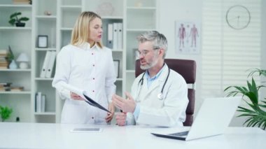 Bir grup doktor modern bir hastane kliniğinde toplantı yapıyor. Kadın bir pediyatrist olgun bir sağlık çalışanına raporunu gösterir ve hastaların tedavisi hakkında danışmanlık yapar.