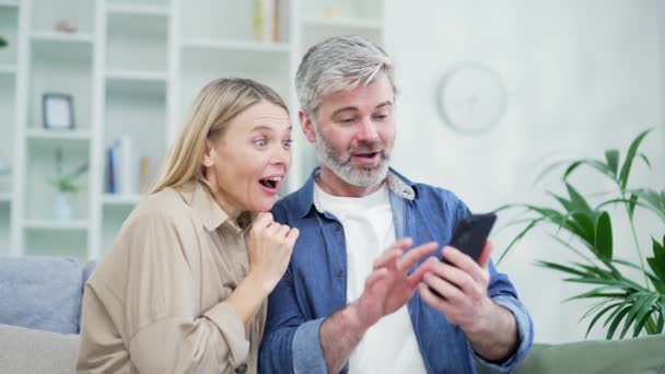 Zbliżenie Zdumiony Szczęśliwy Małżeństwo Sprawdzanie Smartfona Czytanie Wiadomości Podczas Siedzenia — Wideo stockowe