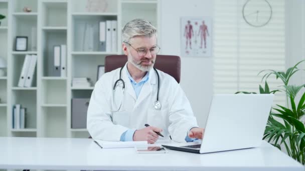 Dojrzały Lekarz Białym Płaszczu Pisze Laptopie Zapisuje Dane Notesie Szpitalnej — Wideo stockowe