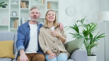 Webcam görüntüsü. Mutlu aile çifti, evdeki oturma odasında kanepede el sallarken görüntülü konuşma yapıyor. Olgun bir eş ve koca kucaklaşır, gülümser, kameraya bakar gibi hareket eder.