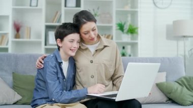Anne ve oğlu birlikte oturma odasında kanepede oturup dizüstü bilgisayarla video izliyorlar, aile birlikte vakit geçirip çevrimiçi eğitim kursları izliyorlar.