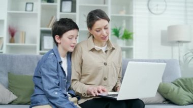 Anne ve oğlu internetten alışveriş yapıyor, dizüstü bilgisayarla internetten alışveriş yapıyor, bir yandan da internette gezinen genç bir sörfçüyle aile kanepesinde oturuyor.