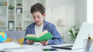 Meraklı okul çocuğu evde bir sınıfta bir masada oturup kitap okuyor. İlköğretim, çocuklar, insanlar, okul konsepti, ödev yazma, bir okul çocuğu ders veriyor.