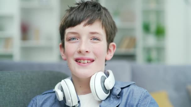 一个脸上挂着雀斑 蓝眼睛 脖子上挂着耳机的男孩快乐地坐在家中房间的沙发上的画像 头像时尚时髦的现代青少年 — 图库视频影像