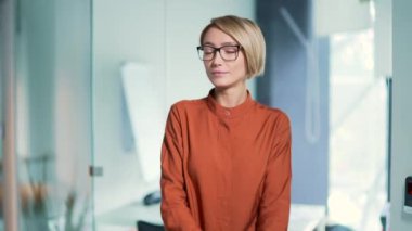 Modern ofiste kollarını kavuşturarak duran kendine güvenen genç bir kadın çalışanın portresi. Gözlüklü şık, ciddi, sarışın bir iş kadını vesikalık iş portresi için poz veriyor. Başarılı bir yönetici