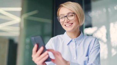 Gözlüklü, gülümseyen genç bir sarışının kapanışı modern bir ofiste otururken akıllı telefondan komik bir mesaj okudu. Neşeli bayan çalışan arkadaşlarıyla sohbet ederken gülüyor, şakalaşıyor.