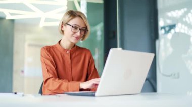 Modern ofisteki iş yerinde otururken dizüstü bilgisayarda daktilo kullanan genç ve mutlu bir çalışan. Gülümseyen sarışın bir kadın internette sohbet ediyor, bir meslektaşıyla, bir arkadaşıyla iletişim kuruyor, bir müşteriye cevap veriyor.