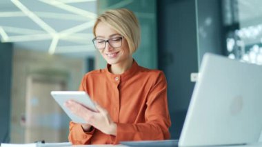 Modern ofisteki çalışma masasında otururken tablet kullanan mutlu genç sarışın kadın. Gözlüklü gülümseyen kadın çalışanlar sosyal ağlara göz atıyor, e-posta mesajlarını okuyor, internette sohbet ediyor.