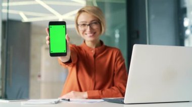Modern ofiste iş yerinde otururken akıllı telefonu yeşil ekranla gösteren genç bir kadın. Renkli anahtar dikey modeli. Reklam şablonu. Mutlu bayan çalışan kameraya bakıyor.