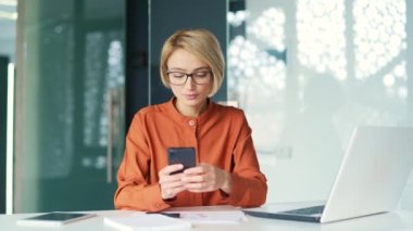 Üzgün genç sarışın kadın, modern ofisteki çalışma masasında otururken akıllı telefondan kötü haberler okuyor. Gözlüklü kadın çalışanın rahatsız edici bir mesaj ya da tebligata bakması.