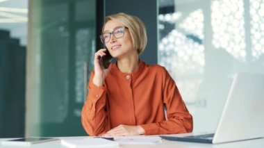 Modern ofisteki çalışma masasında otururken akıllı telefondan konuşan mutlu genç sarışın kadın. Gözlüklü bir iş kadını bir meslektaşıyla iş konuşur ya da bir arkadaşıyla sohbet eder.