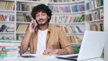 Akıllı telefondan konuşan mutlu üniversite öğrencisi kampüs kütüphanesinde otururken öğretmenine danışıyor. Gülümseyen bir öğretmen meslektaşıyla eğitim süreci hakkında konuşuyor.