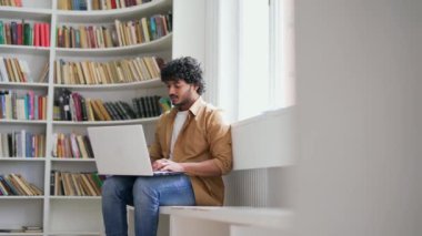 Genç öğrenci, kampüs kütüphanesinde online e-öğrenme için dizüstü bilgisayar kullanıyor. Yakışıklı bir erkek, üniversitenin sınav dönemi için hazırlanıyor. Sınıfta bir bankta oturuyor.