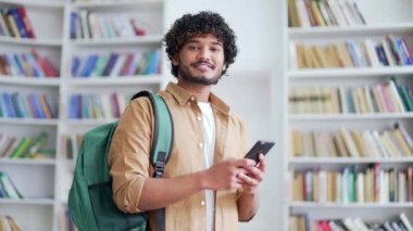 Elinde akıllı telefon olan ve kampüs kütüphanesinde kameraya bakan gülümseyen genç üniversite öğrencisinin portresi. Üniversite sınıfında sırt çantalı, mutlu, arkadaş canlısı bir erkek.