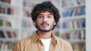 Kampüs kütüphanesinde dikilirken kameraya bakan ciddi bir üniversite öğrencisinin portresini yakından çek. Üniversite sınıfında poz veren gömlekli sakin bir erkek. Yakışıklı, kıvırcık bir esmerin portresi