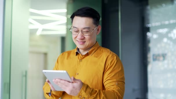 現代のオフィスに立っている間 タブレットを使用して眼鏡でアジアのビジネスマンの笑顔 シャツの閲覧 電子メールのチェック オンラインチャット またはクライアントにメッセージを書くことで幸せな起業家を自信を持って — ストック動画