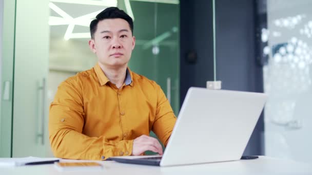 一个亚洲商人坐在现代化办公室的办公桌前使用笔记本电脑的肖像 一位身穿衬衫 面带微笑的商人正看着摄像机 电脑上的员工类型 — 图库视频影像