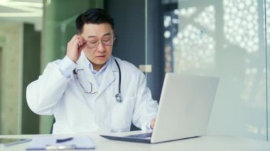 Göz yorgunluğundan muzdarip çok çalışan Asyalı doktor hastane kliniğinde çalışırken dizüstü bilgisayarla çalışıyor. Yorgun bir hekim gözlüğünü çıkarıp şakaklarına masaj yapıyor.