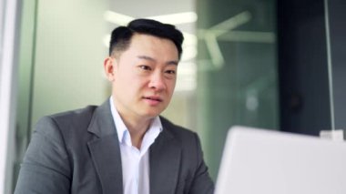 Kapatın. Gülümseyen Asyalı işadamı, ofisteki masasında otururken dizüstü bilgisayarından iyi haberler okuyarak başarıyı kutluyor. Takım elbiseli mutlu adam şok oldu, hoş bir mesaj karşısında şaşırdı.