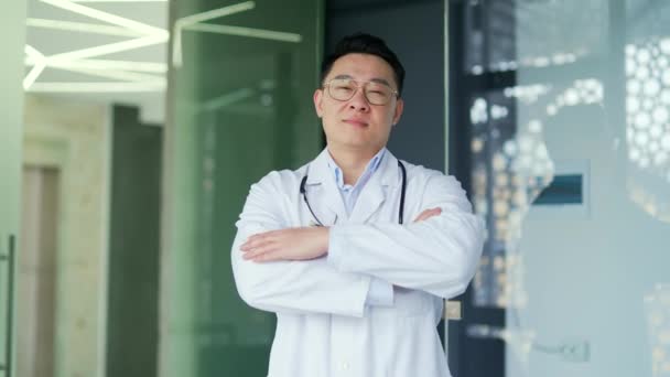 白いコートと眼鏡を着た笑顔のアジア人医師の肖像画は カメラを見て 現代の病院の診療所に立っている 正の医療従事者医師とともに聴診器ポーズで交差腕 — ストック動画