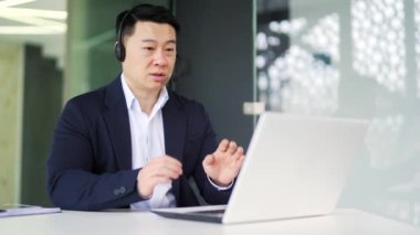 Kablosuz kulaklıklı gülümseyen Asyalı işadamı bilgisayarla video görüşmesi yapıyor, ofiste oturuyor. Takım elbiseli mutlu adam konferansta konuşuyor, iş görüşmesi yapıyor.
