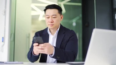 Resmi takım elbiseli ciddi bir Asyalı işadamı ofisteki bir iş yerinde otururken akıllı telefon kullanıyor. Konsantre olmuş düşünceli sahibi istemciye e- posta ya da metin mesajı okuyor, kontrol ediyor