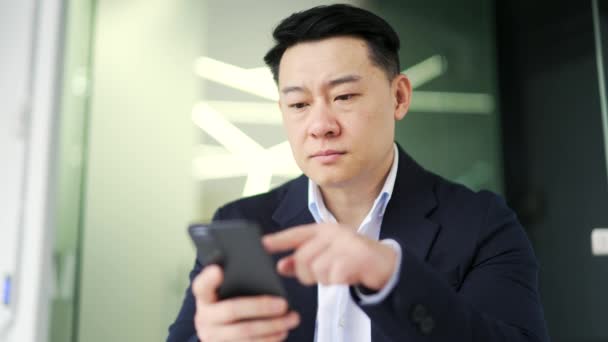 身穿正装的严肃的亚洲商人坐在现代办公室的工作场所时 正在使用智能手机浏览 有识之士 有重点的所有者阅读 检查电子邮件 在线监控市场情况 — 图库视频影像