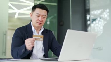 Mutlu Asyalı işadamı, bilgisayar kullanarak online alışveriş yapıyor ve modern ofisteki çalışma masasında otururken kredi kartı numarasını giriyor. Resmi takım elbiseli adam, malların terfisinden memnun.