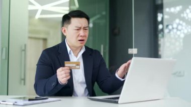Sinirli Asyalı adam, modern ofiste çalışırken kredi kartı numarasını bilgisayarına girerken dolandırıcılığı tespit etti. Endişeli iş adamı, hesabından para çalındığı için üzgün.