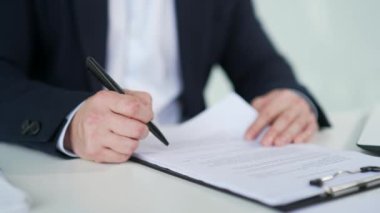 Modern bir ofisin çalışma masasında bir adamın el imzalı belgelerini kapatıyoruz. Resmi takım elbiseli patron dosyaya evraklarla bakıyor ve sözleşmeyi kalemle imzalıyor.