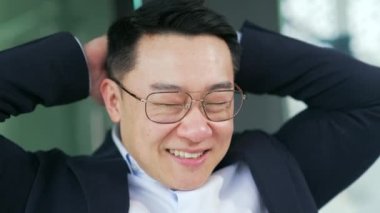 Resmi takım elbiseli ve gözlüklü mutlu Asyalı işadamı modern ofiste çalışırken elleri başının arkasında rahatlıyor. Projeyi bitirdikten sonra gülümseyen girişimci dinleniyor