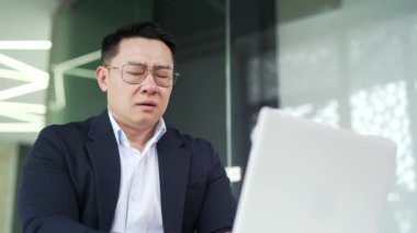 Modern ofisteki çalışma masasında otururken dizüstü bilgisayarla uğraşan yorgun Asyalı adamı kapatın. Resmi takım elbiseli bir iş adamı masaj yapar ve elleriyle şakaklarını ovuşturur.