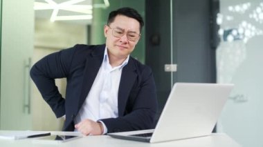 Modern ofis işyerindeki masa başında çalışırken sırt ağrısı çeken Asyalı bir adam resmi takım elbiseli hasta bir çalışan sırt kası spazmları geçirir, sırtını sıvazlar, sırtına masaj yapar,