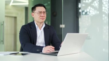 Ofiste çalışırken dizüstü bilgisayarla çalışan Asyalı bir adam işaret parmağıyla dudaklarını işaret ediyor. mployee kulaklarını kapatır, bir sessizlik jesti gösterir. Sessiz olun diyor, sessiz olun diyor.