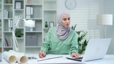 Genç kadın mimar yaratıcı tasarımcı çizimleri olan bir dizüstü bilgisayarı kullanıyor, tarıyor. Ofisteki tesettürlü Arap kadın apartman veya ev inşaatı ve konut yenileme tasarımları yapıyor