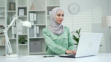 Gülümseyen genç Müslüman çalışan iş kadını modern hafif iş yerinde dizüstü bilgisayarda çalışıyor tesettür işinde çalışan meşgul girişimci işçi harika iş gününün tadını çıkarıyor ve ofisin içinde yazı yazarak arama yapıyor.