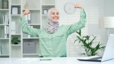 Güçlü, kendine güvenen Müslüman kadın iş adamının portresi kameraya bakıyor ve güçlü bir hareket gösteriyor. İş yerindeki tesettürlü mutlu kadın iç mekan kaslarını gösteriyor.