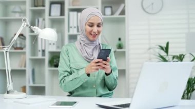 Genç, güzel, Arap iş kadını akıllı telefon yazıcı sohbeti yapıyor. Sörf kayışı yapıyor, cep telefonunu iş yerinde tutuyor. Kapalı alanda çalışan mutlu kadın, uygulama mesajı kullanıyor.