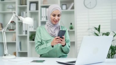 Aşırı neşeli, tesettürlü genç Müslüman iş kadını iş yerindeki Smartphone 'dan mutlu bir şekilde okuyun. Heyecanlı İslami çalışan ekranlara bakıyor ve sevinçten havalara uçuyor.