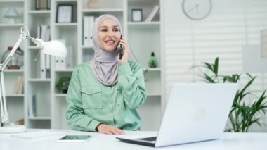 Genç çekici Müslüman iş kadını iş yerinde modern ofiste otururken telefonla konuşuyor. Olumlu gülümseyen Arap İş Kadını Kapalı alanda akıllı telefon kullanarak iletişim kuruyor