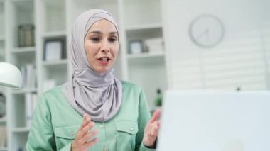 Genç ve güzel bir iş kadını ile tesettürlü Müslüman bir kadın uzaktan haberleşme ve toplantı konferans hoparlörleri için bilgisayarla video üzerinden konuşuyor. Modern bir ofiste ya da ev kapanışında.
