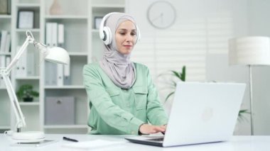 Genç Müslüman kadın çalışan dizüstü bilgisayarda çalışıyor ve podcast dinliyor, müzik dinliyor ya da kulaklıkla radyo dinliyor modern ofiste çalışanlardan memnun kız öğrenci iş yerinde iş yerinde eğleniyor.