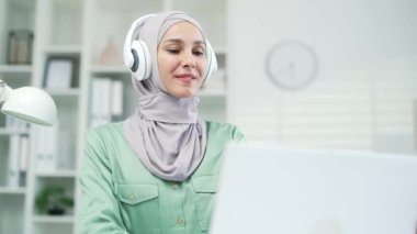 Genç Müslüman kadın çalışan dizüstü bilgisayarda çalışıyor ve podcast dinliyor, müzik dinliyor ya da kulaklıkla radyo dinliyor modern ofiste çalışanlardan memnun kız öğrenci iş yerinde iş yerinde eğleniyor.