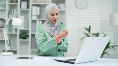 Eklem ağrısı çeken genç bir kadın işçi iş yerindeki dizüstü bilgisayarda çalışırken eline masaj yapıyor. Bileği çok ağrıyor ve uzun süredir klavyede ofis sendromu var.