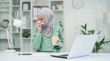 Kadın işçi burnu akan hapşırır. Çalışan soğuk algınlığı ya da grip öksürüyor. Ofiste oturuyor, kendini iyi hissetmiyor. İş yerinde bir bardak sıcak içecekle genç ve güzel müslüman bir kadın.
