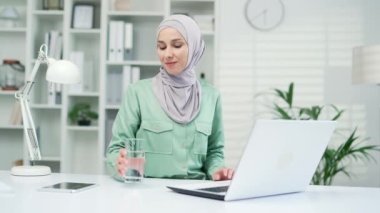 Modern ofisteki dizüstü bilgisayarda çalışırken camdan temiz su içen pozitif Müslüman iş kadını. Gülümseyen girişimci işçi, iş günü kapalı alanda sağlık dengesini koruyor.